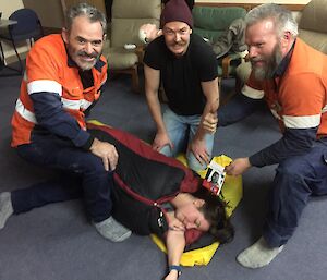 Three expeditioners smiling to camera while working on 1st aid patient (during training) in sleeping bag lying on floor. One expeditioner holds the AAD first aid manual and gives a thumbs up