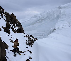 Man in cold weather gear, brown jacket and black beanie, dwarfed when standing in large wind scour around rocky hill’s base