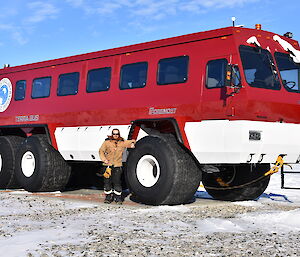 Man stands beside the wheel of a large red bus, wheel comes to shoulder height. Rocky snow covered ground with blue sky above