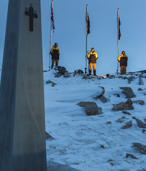 Cenotaph built as stone oblisk with cross at top with poppies at base in foreground, in background on snow covered hill are three flag poles with UK, Australian and NZ flags lit up in the dawn light, with an expeditioner standing at the base of each flag pole