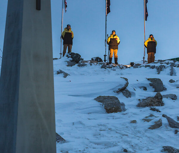 Cenotaph built as stone oblisk with cross at top with poppies at base in foreground, in background on snow covered hill are three flag poles with UK, Australian and NZ flags lit up in the dawn light, with an expeditioner standing at the base of each flag pole