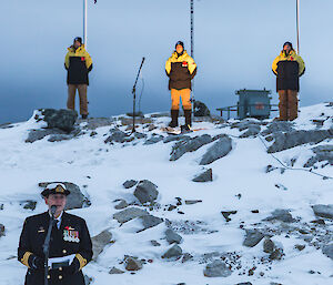 Woman in navy uniform in front left, behind on hill are the three flag poles with three expeditioners at their base