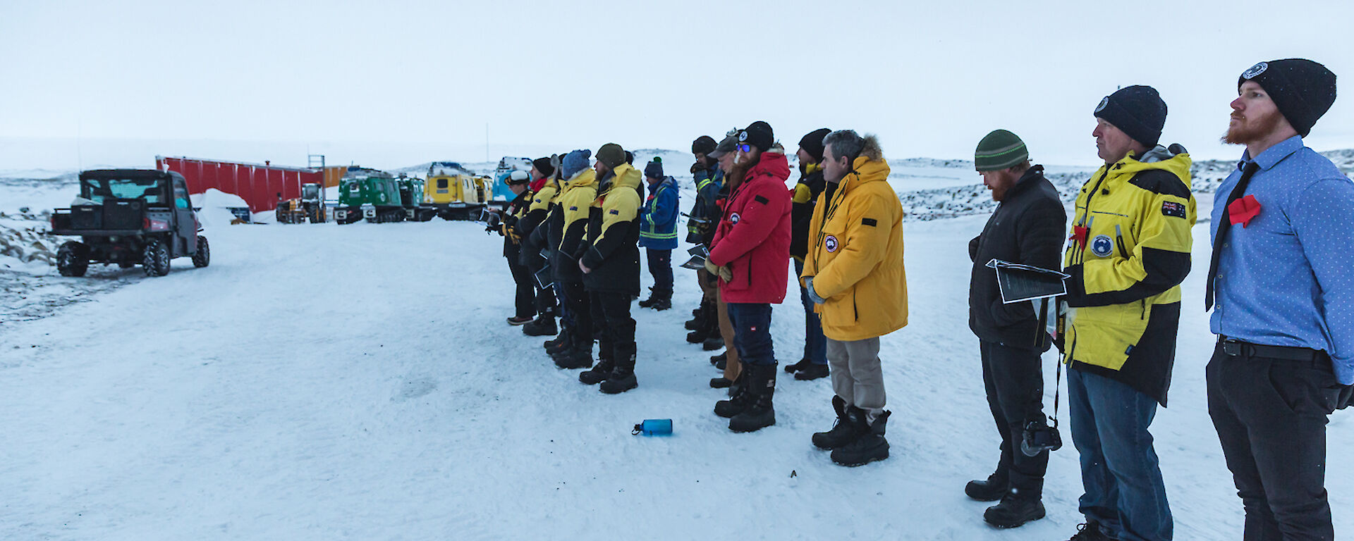 Group of expeditioners standing with bowed heads on snow covered ground looking towards the sunrise. In the background are parked hagglunds vehicles