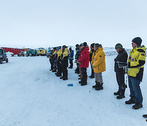 Group of expeditioners standing with bowed heads on snow covered ground looking towards the sunrise. In the background are parked hagglunds vehicles