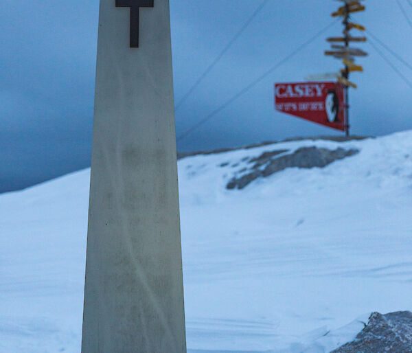Cement look pillar with brass cross at top and group of red poppies at base. In the background the Casey distance to sign on snow covered hill