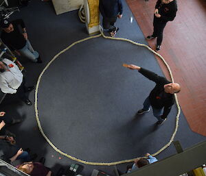 Looking down from above on man standing inside rope circle, tossing two coins into air from wood paddle. Expeditioners stand around the circle looking on