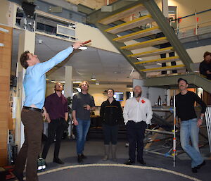 Two up underway, man to left throws coins in the air with wooden paddle inside rope circle. Expeditioners look on from outside the circle and sitting on stairs going up to first floor