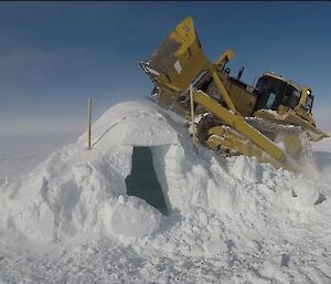 Ice igloo with door facing photographer being crushed by bulldozer which has driven up onto it’s roof