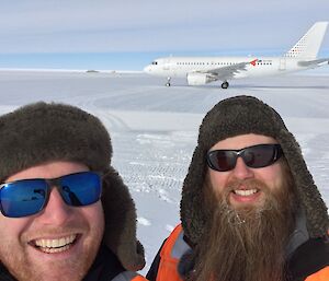 Head shot of two expeditioners in woolen hats and sunglasses with ice runway and white A319 plane in background