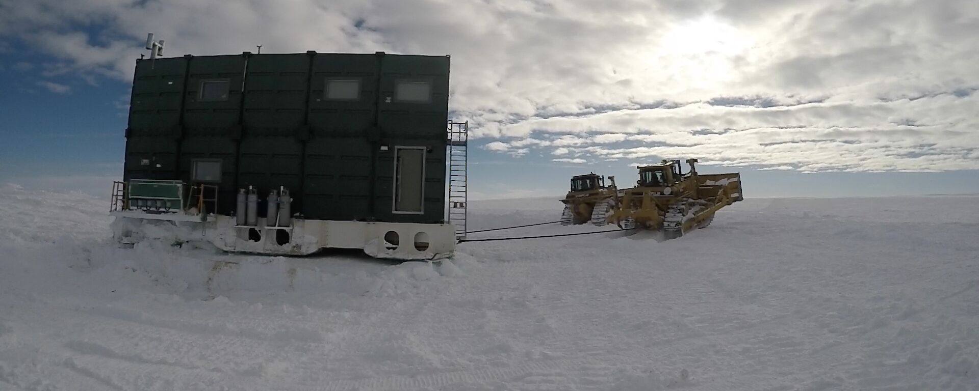Large green building on skids being towed by two tractors across the snow