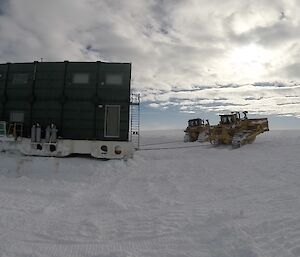 Large green building on skids being towed by two tractors across the snow