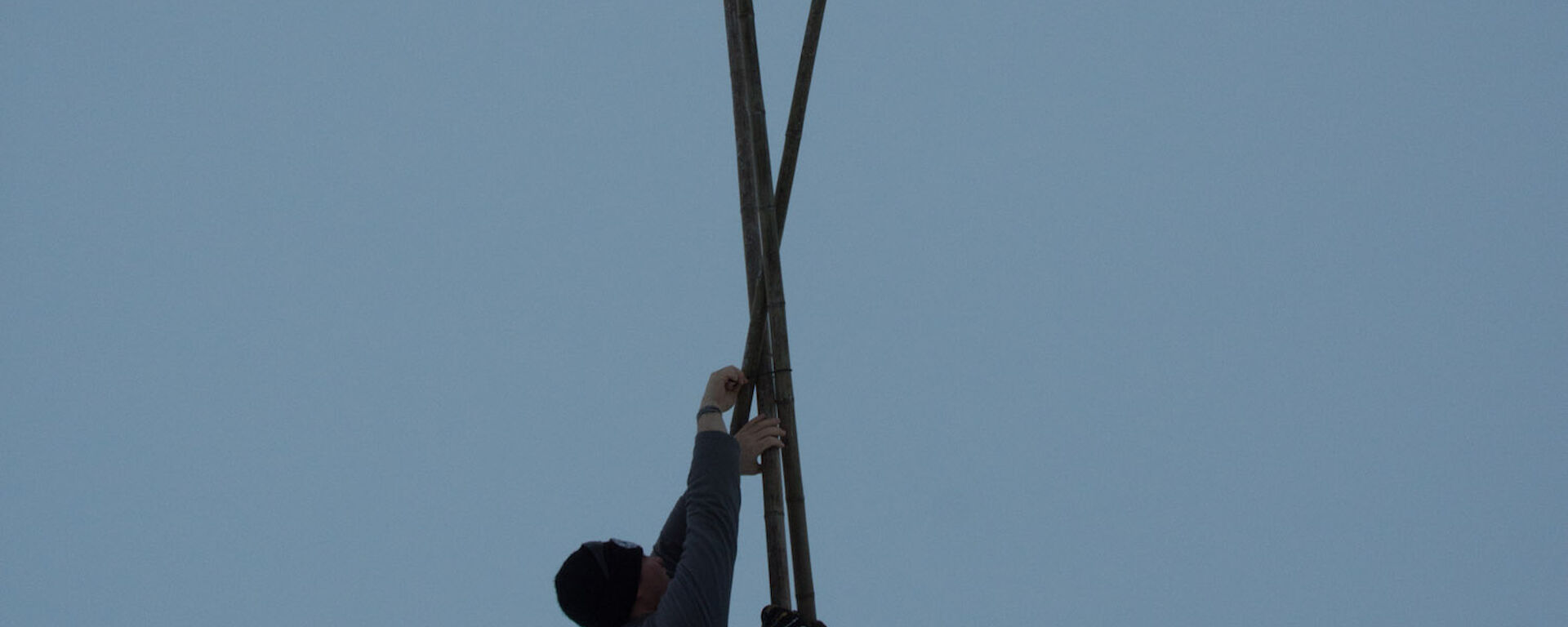 Three canes with flouro orange flags at top set up in teepee shape, with man and woman either side reaching up above heads to afix point where the canes join. Ice drill standing on end in left foreground