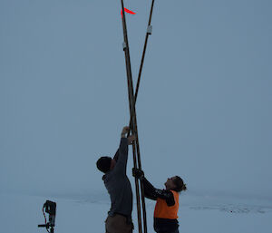 Three canes with flouro orange flags at top set up in teepee shape, with man and woman either side reaching up above heads to afix point where the canes join. Ice drill standing on end in left foreground