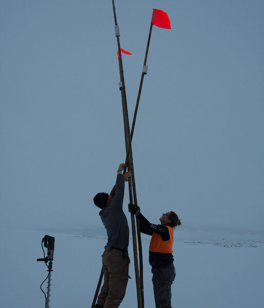 Three canes with flouro orange flags at top set up in teepee shape, with man and woman either side reaching up above heads to afix point where the canes join. Ice drill standing on end in left foreground