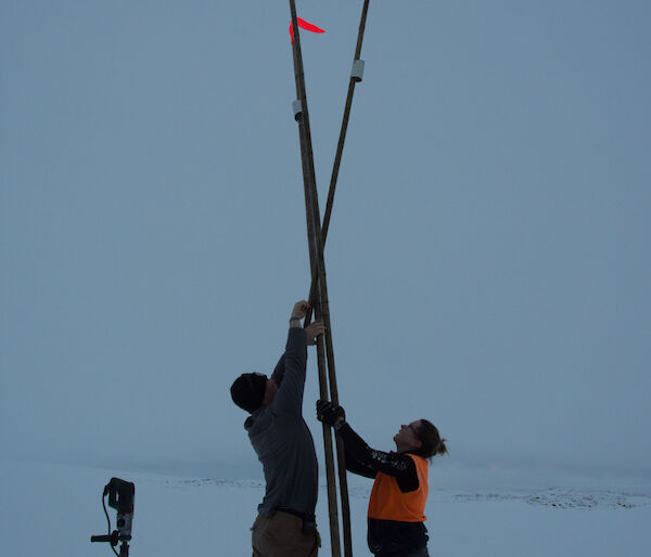 Three canes with flouro orange flags at top set up in teepee shape, with man and woman either side reaching up above heads to afix point where the canes join. Ice drill standing on end in left foreground