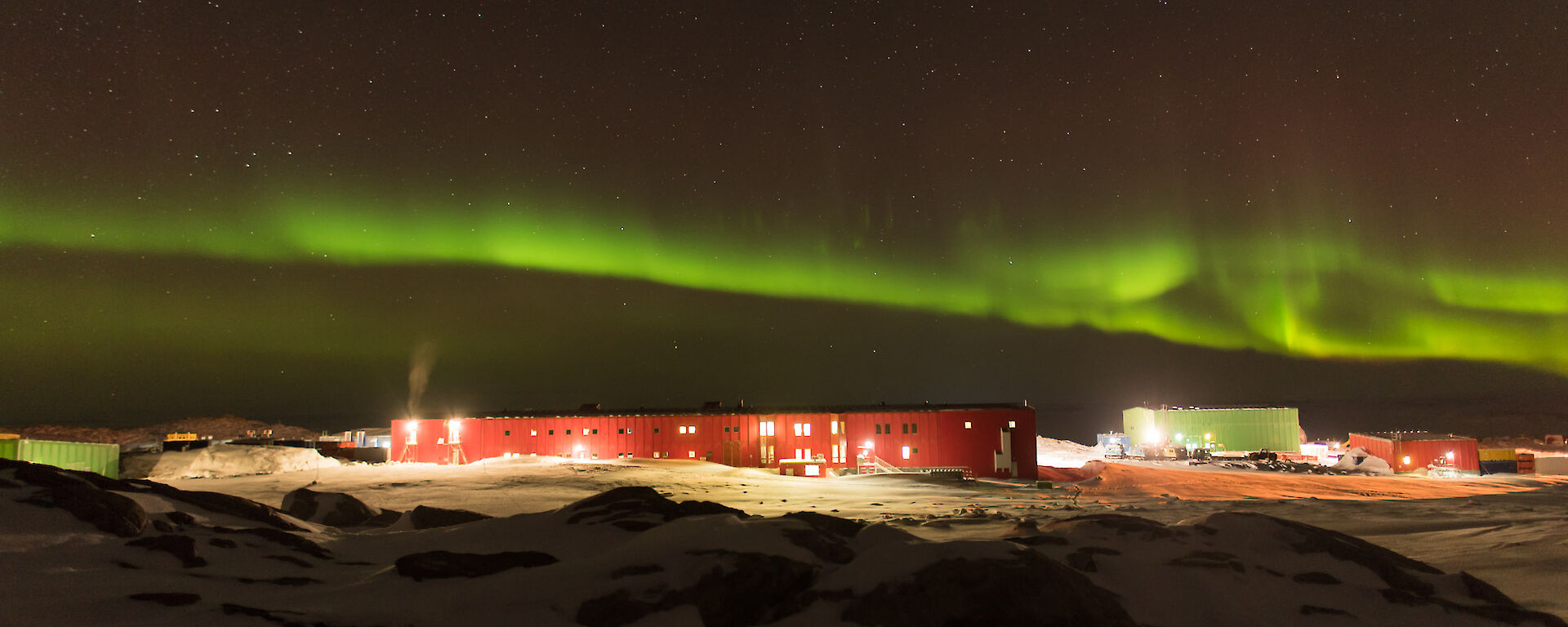 Mid picture the station in distance lit up by lights at night and above a bright green band of aurora australis / southern lights