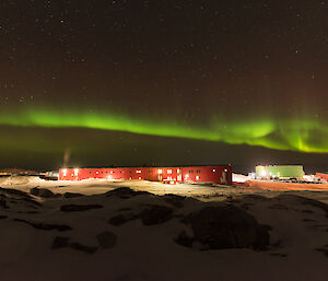 Mid picture the station in distance lit up by lights at night and above a bright green band of aurora australis / southern lights