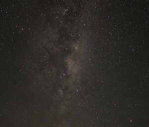 Night sky fills the picture with bright stars and the fog of the milky way, in foreground is the sillouette of a photographer standing at tripod looking through camera’s viewfinder