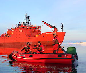Three men in red and black dry suits in inflatable boat in foreground, behind is the Aurora Australis (large orange icebreaker ship).