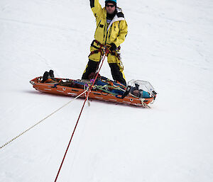 Expeditioner roped up and down icy slope stands over person in a stretcher