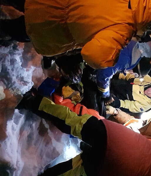 Man lying down on snow with group of people gathered around, In the dark and faces of men highlighted by torches, held and on heads
