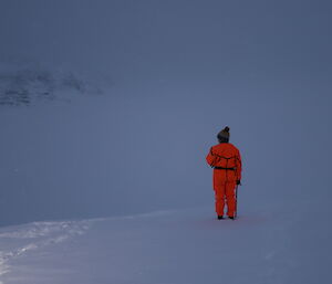 Woman standing with back to camera, in flouro orange dry suit and beanie with pom-pom. All around is white, snow covered ground blending into foggy sky