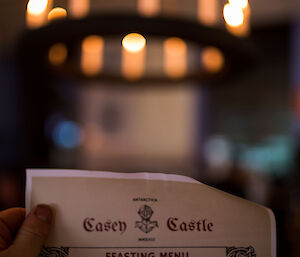 Bottom half of picture, the feasting menu held up by one hand on left, above in top of picture a candle laden round wooden chandelier attached to roof by black chains