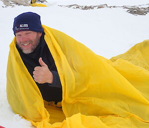 Man giving the thumbs up as he sticks head out of bright yellow bivvy bag stretched out on the snow covered ground