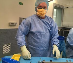 Man dressed in surgical scrubs with gloves and mask staning in operating theatre in front of tray of instruments