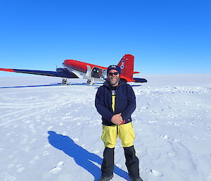 Man in outdoor gear standing centre foreground, in the background a red and white DC3 Basler aircraft on ice runway, bright blue skies above