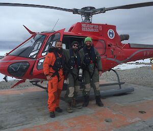 3 men standing in front of a red helicopter, parked on a concrete helipad with snow covered ground in background