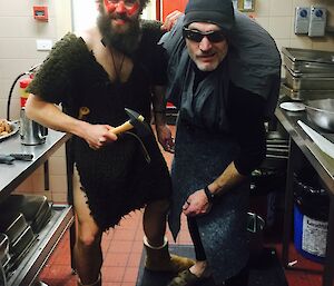 Two men posing in kitchen, man on left dressed as a barbarian with red painted face and fur tunic, and man of right dressed as a hunchback with grey sheet over pillow on back wearing grey beanie and sunglasses