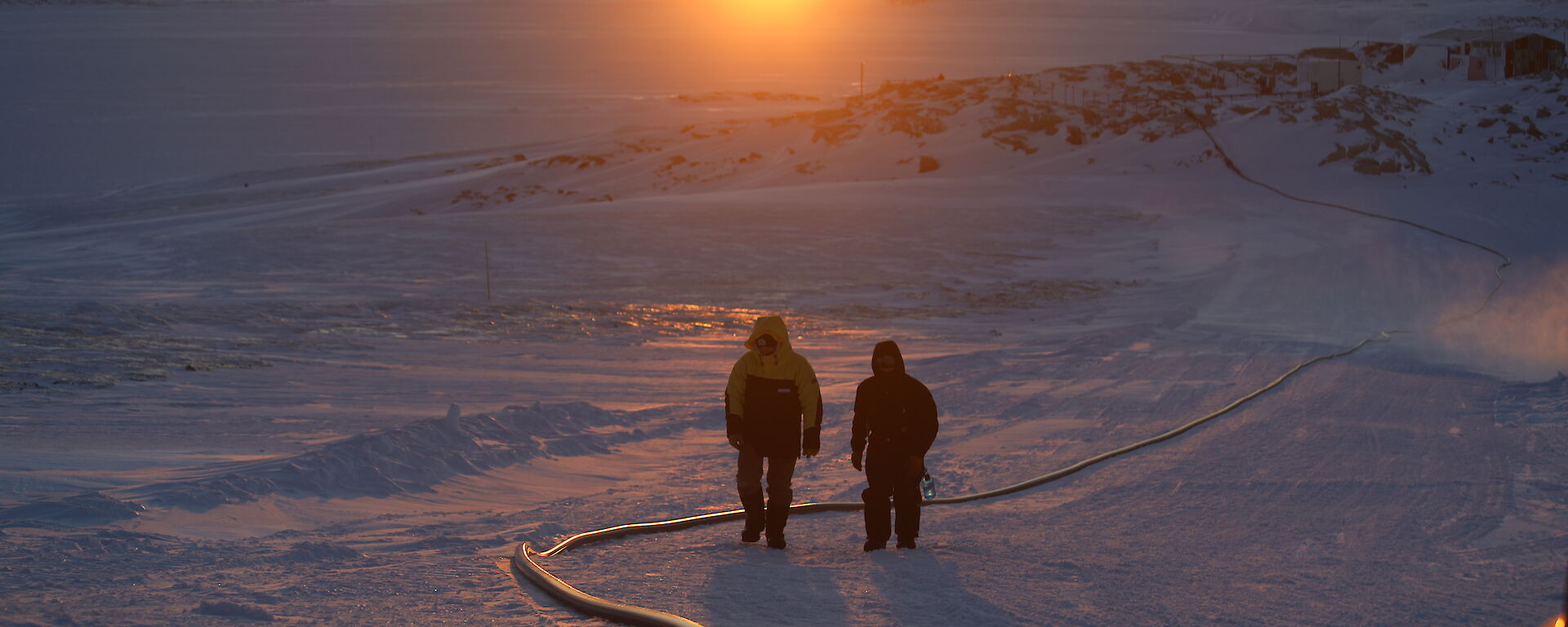 Two men walking centre picture along a large fuel hose which is lying along the snow, in the distance rocky ridges with the sun just rising on the horizen