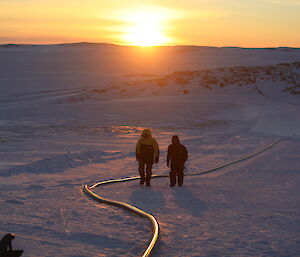 Two men walking centre picture along a large fuel hose which is lying along the snow, in the distance rocky ridges with the sun just rising on the horizen