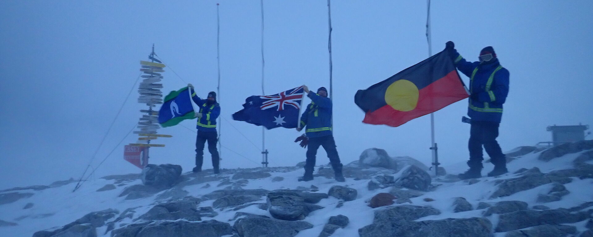 Men standing holding the Torres St Islander flay, Australian National Flag, and Aboriginal Flag in the strong wind. Standing on rocky outcrop with three flag poles and casey sign behind