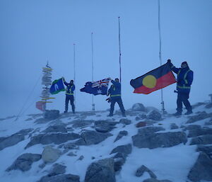 Men standing holding the Torres St Islander flay, Australian National Flag, and Aboriginal Flag in the strong wind. Standing on rocky outcrop with three flag poles and casey sign behind