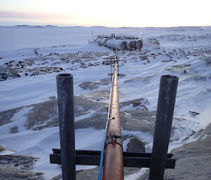 Looking down a steel pipe towards large fuel tanks, pipe extends across snow covered rocky ground, in the distance is an ice covered bay