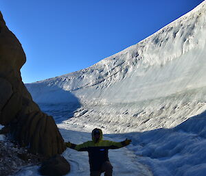 Expeditioner in foreground on knees holding arms out, behind is large icy wind scour dominating the piture and wrapping around a rocky slope to left of picture