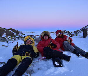 Three men sittin gin the snow, drinking a beer, with sunset behind