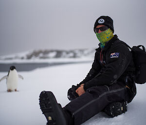 Man sitting in foreground to right of picture, to distant left is small adelie penguin walking towards man, in the background is icy landscape with grey water in bay