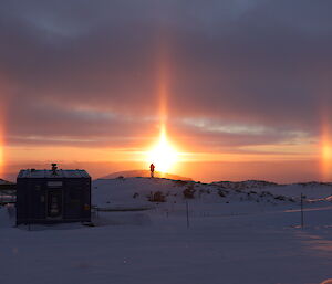 Sun is rising and forming sun dogs, in the foreground a man is on small hill and sillouetted against the sun
