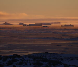 Distant icebergs on the far horizen lit up with orange and purple light at sunrise, steam fog coming from areas of open water, sea ice forming in the closer water and foreground is rocky ground