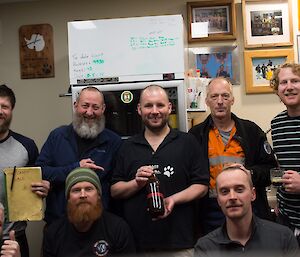 Group of men in brew room with centre man holding a brown beer bottle with red tape reading 10000 on it