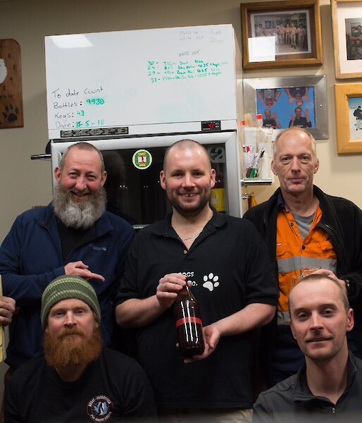 Group of men in brew room with centre man holding a brown beer bottle with red tape reading 10000 on it