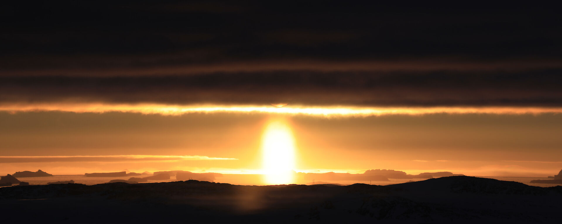 Panoramic shot of the distant horizon with dark foreground and dark clouds above and then sun in centre glowing brightly and sending golden light across the centre of the picture