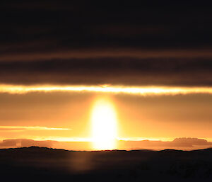 Panoramic shot of the distant horizon with dark foreground and dark clouds above and then sun in centre glowing brightly and sending golden light across the centre of the picture