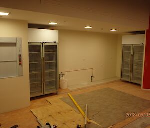 Back end of large room showing L to R: dumb waiter lift doors, double doored fridge, blank wall for servery area, 2nd double doored fridge, and partial wall with rough orange paint. Floor infront is pale orange lino with work tools scattered across