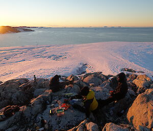 Men sitting around a cheese platter on rocky outcrop in foreground, in background is a stretch of sea ice then open water with the sun setting