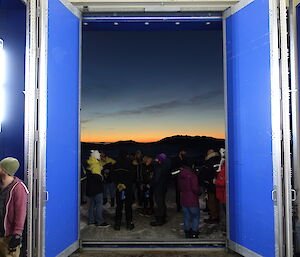 Photo taken from inside blue building with double width and heigh doors opened inwards and group of people gathered outside. In the distance the sun is setting over hills sillouhetted against night sky