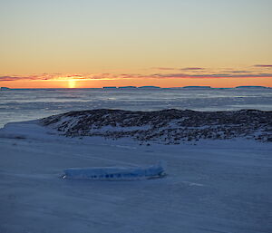 Sea ice with trapped iceberg in foreground, then rocky island middle, with open water other side of island extending to horizen where the sun is setting